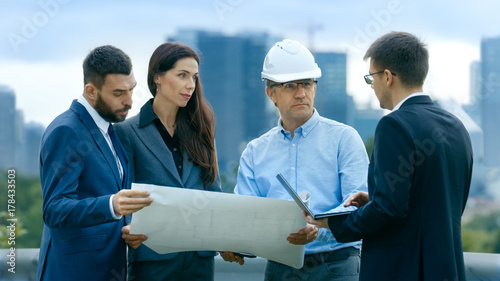 Team of Investors, Developers and Engineers Discuss Future of the Major Real Estate Project with Major Contractor while Consulting Drafts and Plans. In the Background Big City with Skyscrapers.