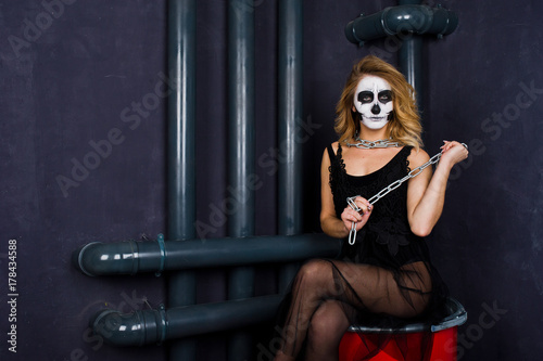 Halloween skull make up girl wear in black against sitting on red barrel at studio.