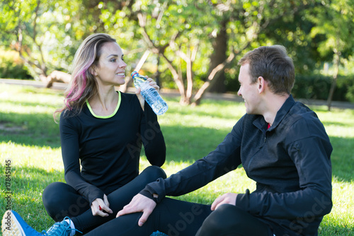 Young happy sporty couple sitting on a meadow in a park after training, drink water © Oleg