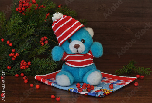 A small toy teddi bear in a red hat andspruce branches with red berries  next to it on an old wood background. Christmas and New Year cards. Still life. photo