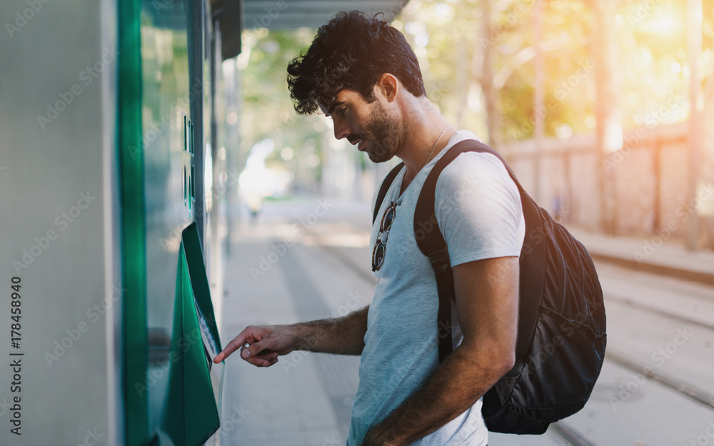 Bearded handsome hipster guy wearing casual clothes with backpack is buying bus tickets at self service vending machine on the station during vacation trip to European city. Flare light. - obrazy, fototapety, plakaty 