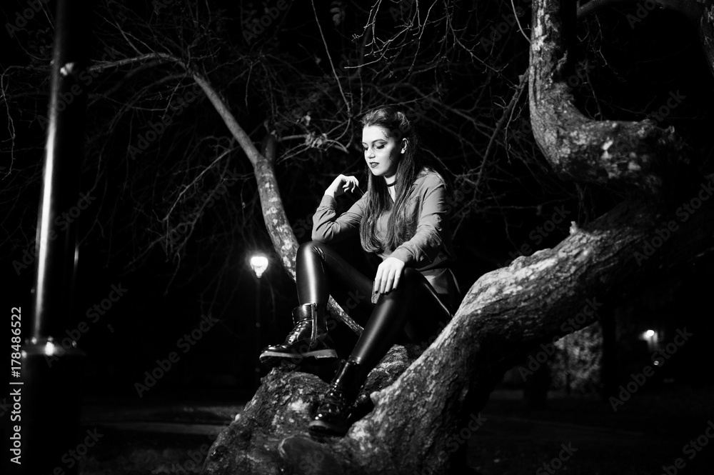Obraz premium Black girl in red shirt and bright make-up on night street sitting on scary tree outdoor. Halloween theme.