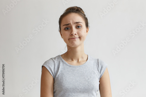 Portrait of upset hopeless young female with blue eyes and hair knot frowning eyebrows and looking at camera with disappointed smile  her look expressing sorrow as if she sas  It is what it is