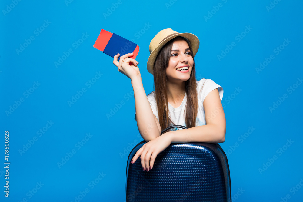 Portrait of trendy young female in wicker hat standing with suitcase and holding passport with tickets, over blue background Portrait of trendy young female in wicker hat standing with suitcase and holding passport with tickets, over blue background