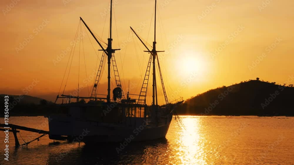 Small yacht moored in calm water near the shore on sunset, sun setting behind hill Small yacht moored in calm water near the shore on sunset, sun setting behind hill
