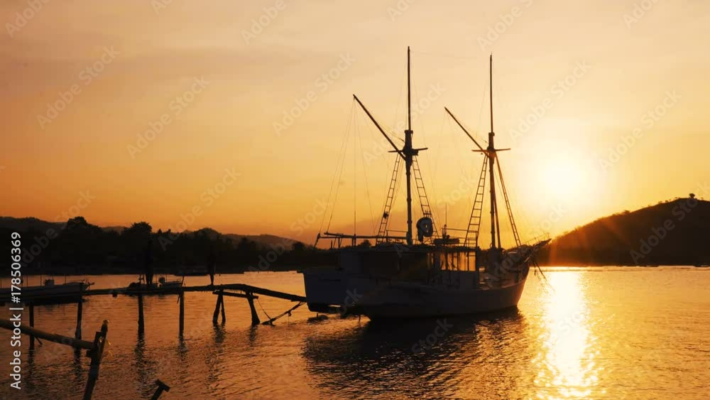 Small yacht moored in calm water near the shore on sunset, sun setting behind hill Small yacht moored in calm water near the shore on sunset, sun setting behind hill