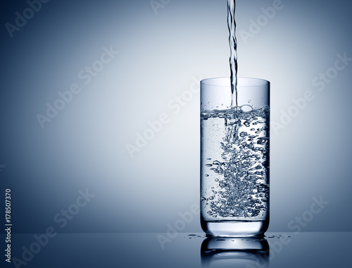 Water pouring and splashing into glass with reflection on blue grey background