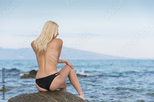 Billede på lærred Slim gorgeous blonde woman sitting in rear view topless on the rock gazing the s