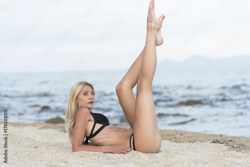 Slim tall gorgeous blonde woman at the beach lying on sand lifting her long legs