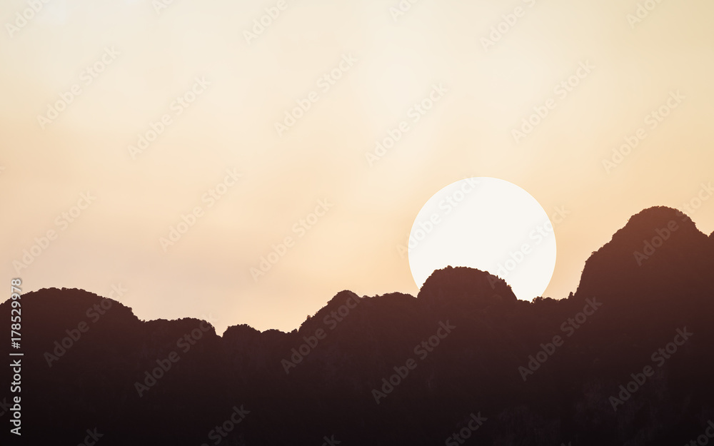 Sunset scene of moving sun over the edge of silhouetted dark mountains range with clear orange warm sky.