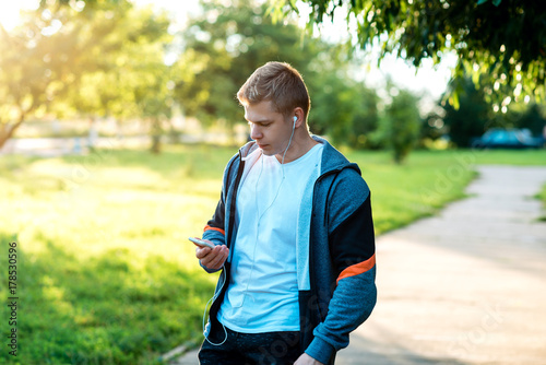 A man is an athlete in nature, a street in the city, Writing a message in social networks. In the summer in the park. Life style. A smartphone in the hands of listening to music. © byswat