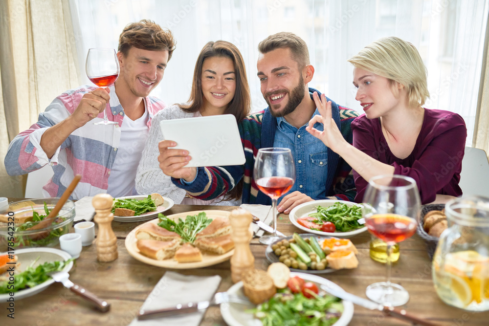 Portrait of four happy people laughing while video chatting from digital tablet at dinner table during holiday celebration