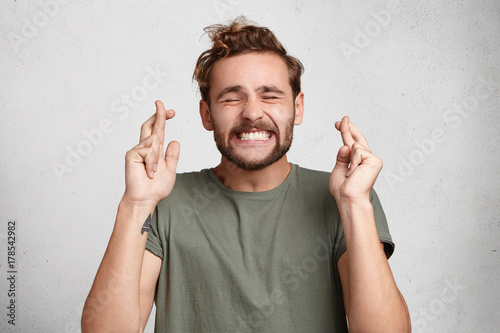 Cheerful bearded man keeps fingers crossed, smiles broadly and closes eyes, makes desire wish. Positive male student believes in victory or success on exam. Feelings, attitudes and reactions photo