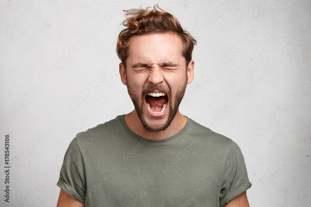 Emotional youngster yells loudly, opens mouth broadly, feels desperate after finding out about tragic events, poses against white studio wall. Caucasian bearded guy scremas in terror of something Emotional youngster yells loudly, opens mouth broadly, feels desperate after finding out about tragic events, poses against white studio wall. Caucasian bearded guy scremas in terror of something