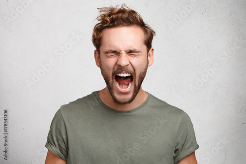 Emotional youngster yells loudly, opens mouth broadly, feels desperate after finding out about tragic events, poses against white studio wall. Caucasian bearded guy scremas in terror of something