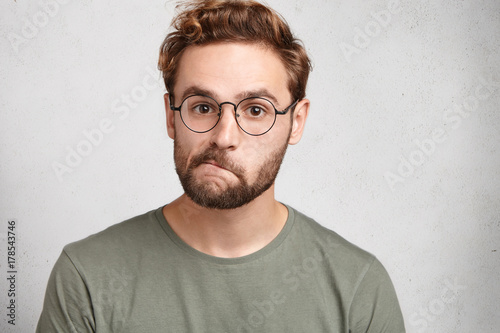 Canvastavla Indoor shot of doubtful bearded man in spectacles, curves lips, looks directly into camera, hesitates what answer to give