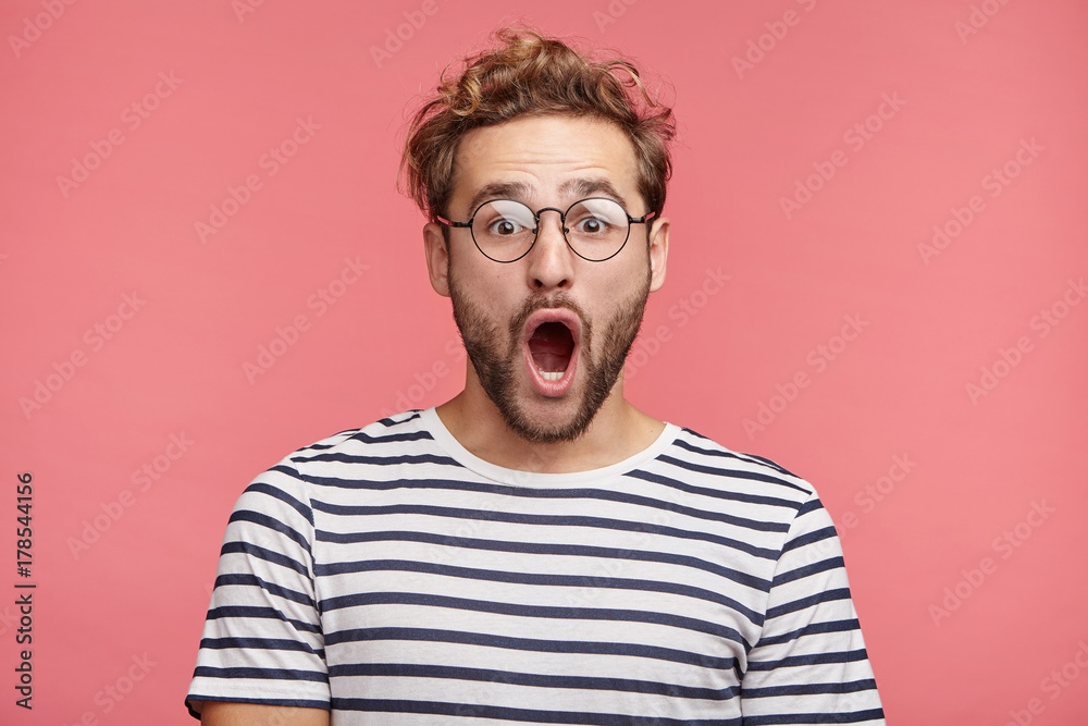 Indoor shot of stupefied handsome male opens mouth widely, wears striped t shirt, has shocked expression, being surprised, isolated over pink background. Astonished young crazy man feels stress Indoor shot of stupefied handsome male opens mouth widely, wears striped t shirt, has shocked expression, being surprised, isolated over pink background. Astonished young crazy man feels stress