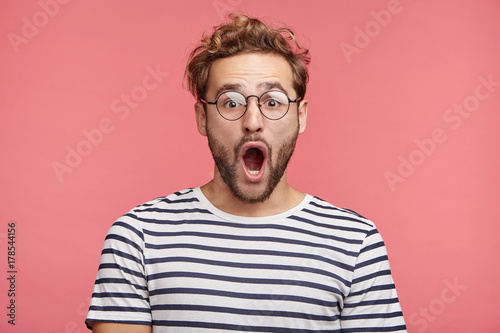 Indoor shot of stupefied handsome male opens mouth widely, wears striped t shirt, has shocked expression, being surprised, isolated over pink background. Astonished young crazy man feels stress © Wayhome Studio