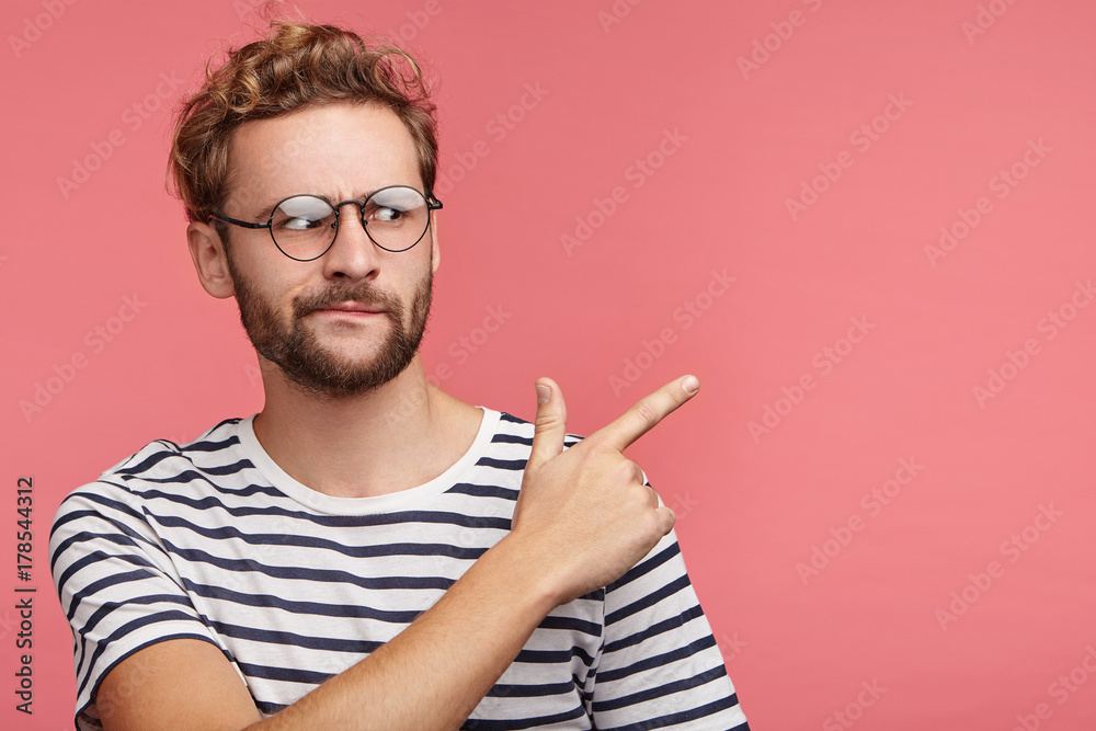 Displeased discontent bearded young man frowns face, indicates at copy space, wears round spectaces and striped t shirt. Puzzled hipster guy looks in bewilderment as sees something unexpected Displeased discontent bearded young man frowns face, indicates at copy space, wears round spectaces and striped t shirt. Puzzled hipster guy looks in bewilderment as sees something unexpected