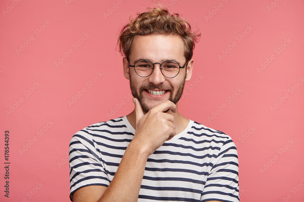 Fashionable stylish male has pleased expression, keeps hand under chin, being happy to recieve congratulation from colleagues. Smiling bearded boyfriend looks at his lover, admires her beauty