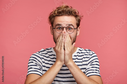 Alarmed nervous hipster guy covers mouth with hands  being shocked or surprised to see unexpected gift prepared by girlfriend. Astonished emotional fearful handsome young male over pink background