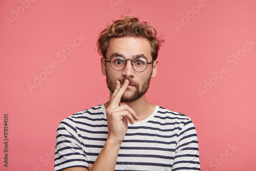 Headshot of stylish man with trendy hairdo, wears round spectacles, keeps fingers on lips, has shocked expression, don`t expect to see his companion at home, didn`t wait for guests, not prepared