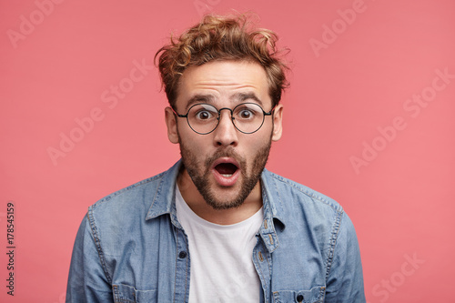 Stupefied attractive young bearded man looks with astonishment into camera, being amazed with negative news. Emotional hipster guy expresses surprisment, doesn`t believe his eyes. Facial expression photo