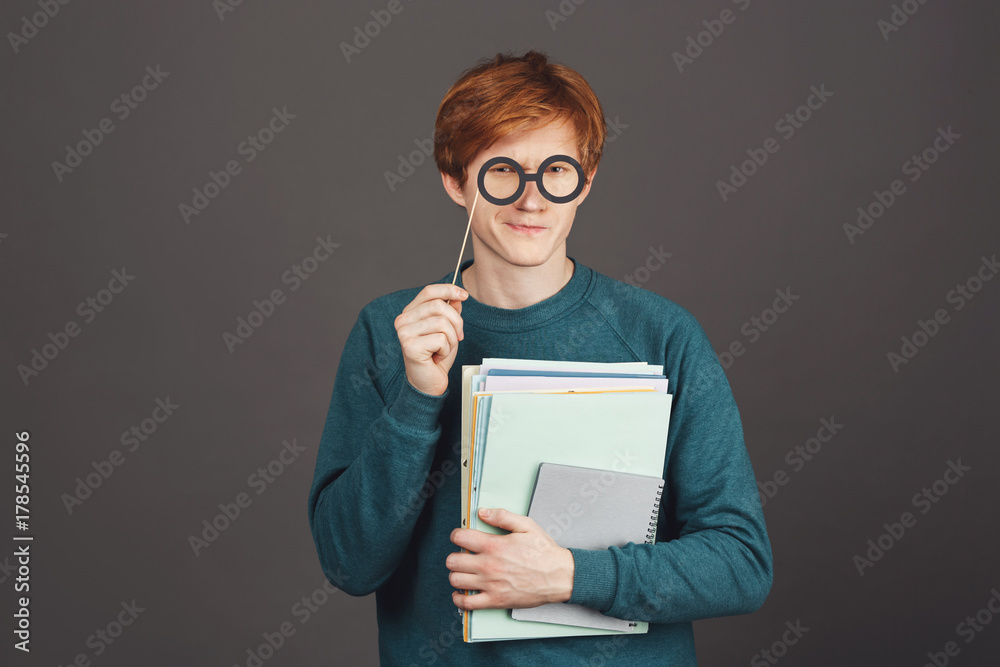 Close up portrait of funny young good-looking ginger guy in green sweater holding lot of notebooks in hand, looking with unsure expression through paper party glasses.