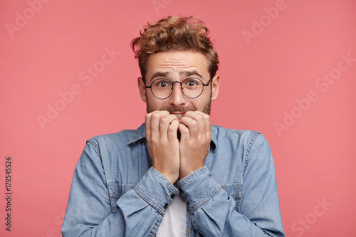 Worried hipster guy bites nails, looks nervous before passing exam or important event in his life. Embarrassed fashionable young man being afraid of difficulties, stands against pink background photo