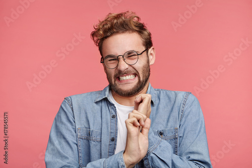 Stylish young male clenches teeth, has digusting expression, refuses do something, makes grimace, isolated over pink background. Dissatisfied youngster in glasses feels averse to do something