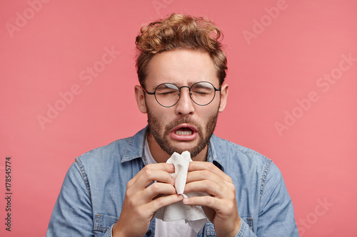 Portrait of ill or sick bearded male sneezes and coughs, uses handkerchief, rubs nose, being allergic to animals. Man has running nose, caught cold when had walk outdoors in winter or autumn photo