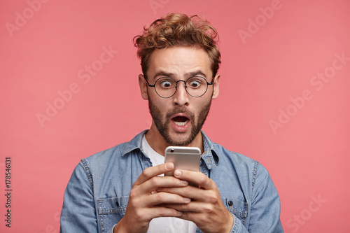 Emotional bug eyed handsome man has trendy hairdo, holds modern mobile phone, being shocked to recieve message from partner, completely forgets about their meeting, has no time for preparation photo