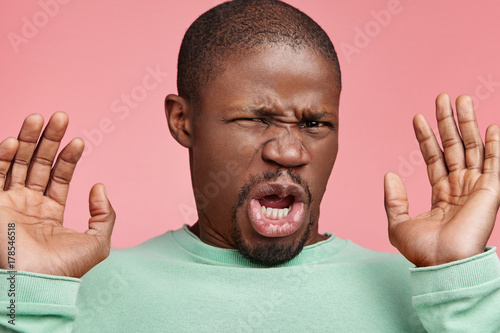 Disgusted African American male feels aversion towards something, frowns face, gestures with hands, has squeamish expression, isolated over pink studio background. Emotional black young adult
