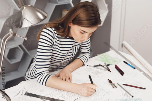 Cropped view of young uropean freelance engineer wearing non-formal striped clothes, sitting at table in comfy coworking space, doing her work, using lot of stationaries.
