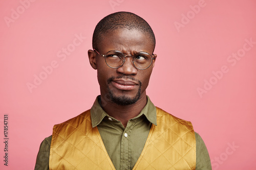 Hesitant African male frowns face in bewilderment, looks pensively aside, tries to recollect something in his mind. Serious dark skinned male freelancer dressed formally, poses against pink wall photo