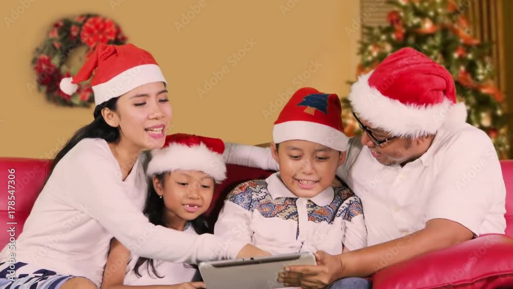 Happy family using a digital tablet computer together on Christmas day while wearing Santa hat and sitting on the sofa with Christmas tree background at home Happy family using a digital tablet computer together on Christmas day while wearing Santa hat and sitting on the sofa with Christmas tree background at home