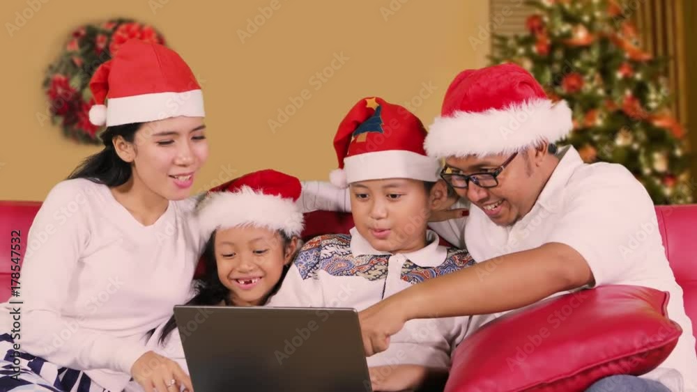 Happy family using a laptop computer together on Christmas day while wearing Santa hat on the sofa with Christmas tree background at home Happy family using a laptop computer together on Christmas day while wearing Santa hat on the sofa with Christmas tree background at home