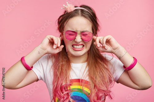 Young frustrated stylish female likes pink colour  blocks ears as hears loud music on party  has headache  clenches teeth  wants silence  isolated over pink background. Cute lovely girl plugs ears