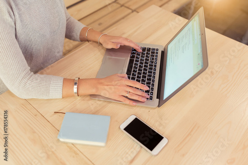 Rear view of young woman using laptop for surfing and browsing Internet, searching for required information, sitting at desk with mobile phone and notebook