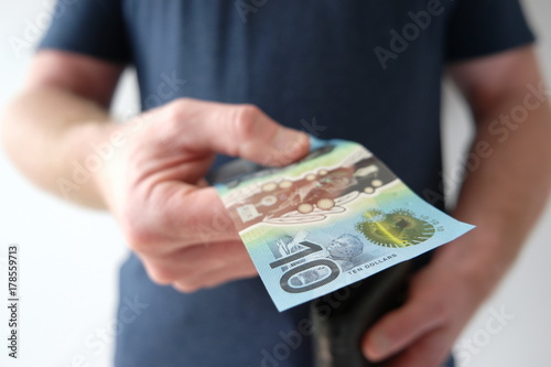 A man holding out a ten dollar Australian currency to buy, donate, share, pay someone, or invest. What does ten dollars get you?