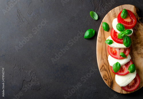 Sliced tomatoes and mozzarella on a wooden board made of olive tree, dark stone background. Top view, copyspace