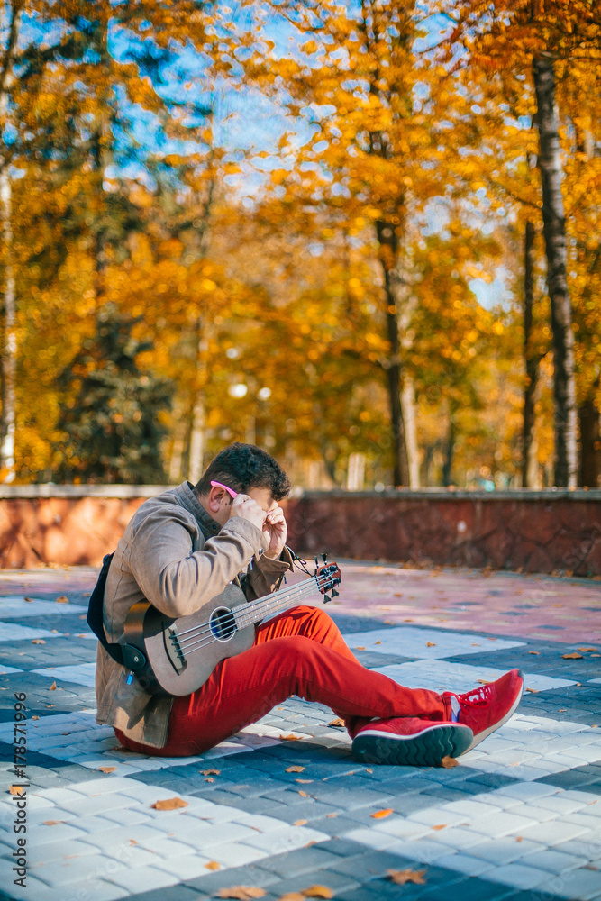 The young musician plays bass ukulele on the street. Man sitting on the ground with guitar. Concept. Basking The young musician plays bass ukulele on the street. Man sitting on the ground with guitar. Concept. Basking