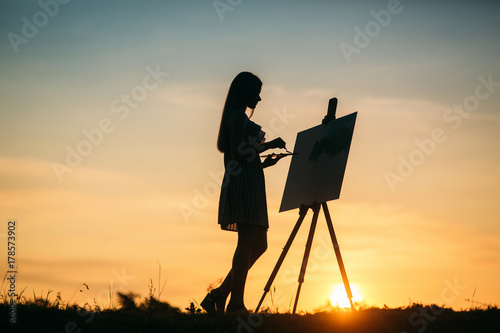 Silhouette of blonde girl paints a painting on the canvas.Palette with paints and spatula, summer sunny day