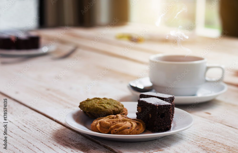 chocolate cookies and brownie in white plate on wood table with coffee cup at morning with sunrise. dessert and coffee concept. chocolate cookies and brownie in white plate on wood table with coffee cup at morning with sunrise. dessert and coffee concept.