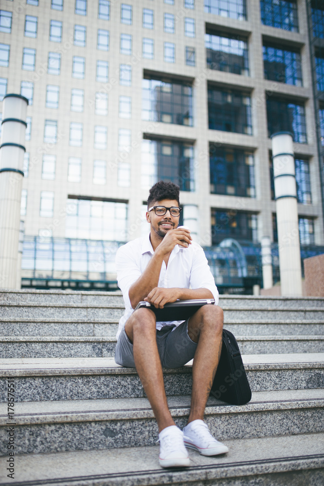 Young handsome Afro American man sitting in front of huge modern business building Young handsome Afro American man sitting in front of huge modern business building