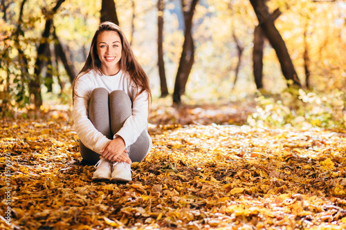 Young gorgeous woman sitting on leaves after warm up and exercises at park or forest. Attractive girl relaxing before fitness in the autumn park.