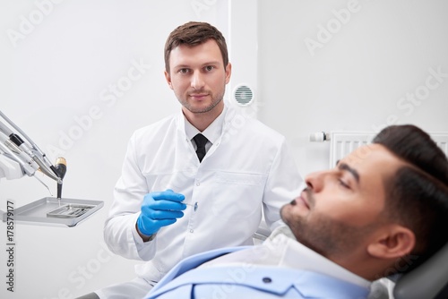 Mature male dentist looking to the camera confidently while working with his patient dentistry oral checkup doctor profession experience occupation healthcare medicine concept.