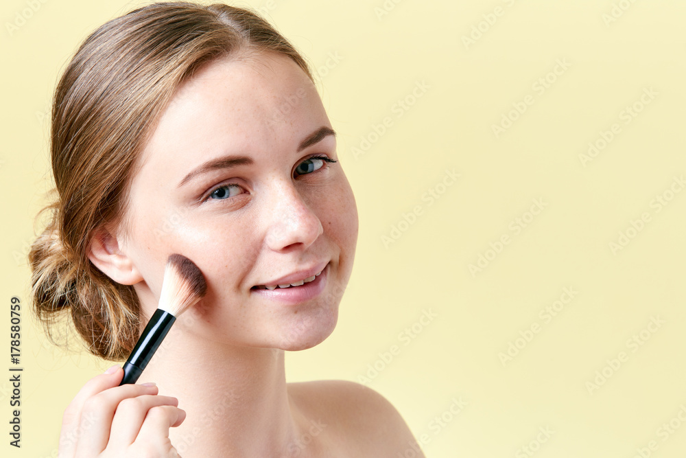 Beautiful young redhead woman with freckles contouring her cheekbones with make up brush. Beauty portrait on yellow background. Beautiful young redhead woman with freckles contouring her cheekbones with make up brush. Beauty portrait on yellow background.