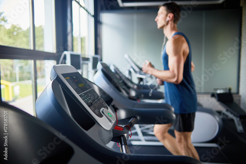 Side view of young man running on treadmill in modern gym by window, focus in foreground on row of machines
