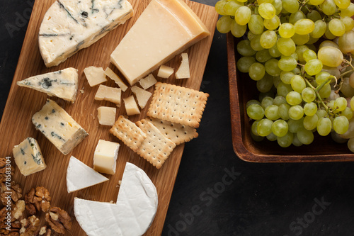 Tasting cheese dish on a wooden plate. Food for wine and romantic, cheese delicatessen on a dark stone table. Top view